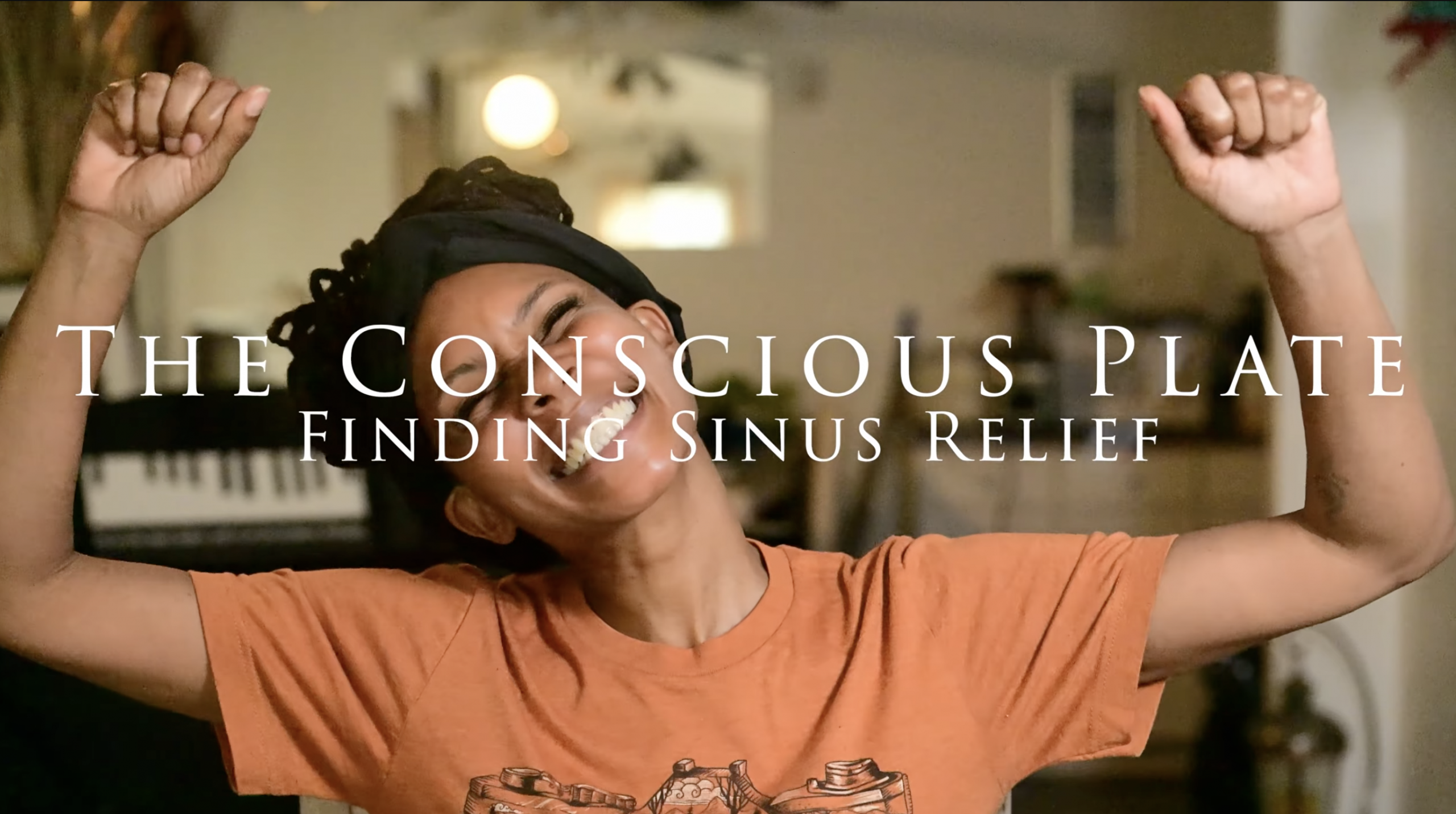 Nikki Brianne speaking directly to the camera, wearing a black scarf and orange t-shirt, introducing the pilot episode of Conscious Plate.